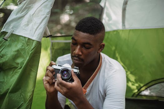 Thoughtful African American male tourist checking old fashioned photo camera sitting in tent during journey in nature