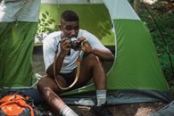 Serious African American tourist resting in tent while using old photo camera in green woods