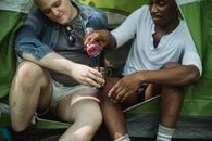 African American male traveler pouring tea into cup from thermos while resting in tent during journey with friend