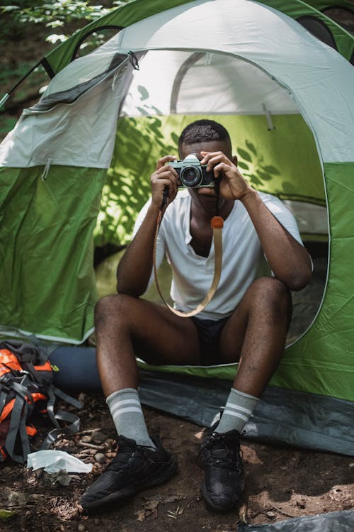Full body of African American camper sitting in tent and taking picture on retro photo camera while traveling across nature