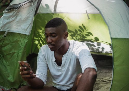 Calm African American male camper sitting in tent and scrolling mobile phone while trekking in nature