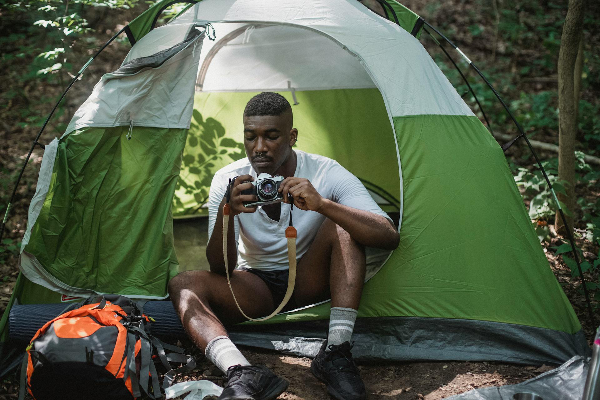 Serious African American male tourist sitting in camping tent with vintage photo camera while hiking in forest