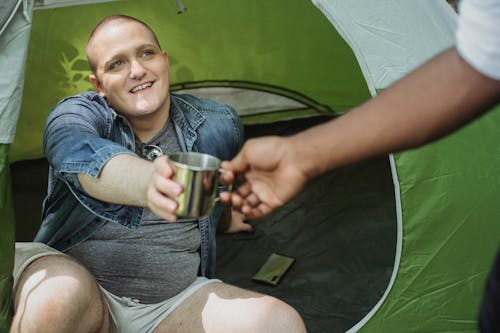 From above of smiling camper giving cup of tea to African American male friend while resting in tent