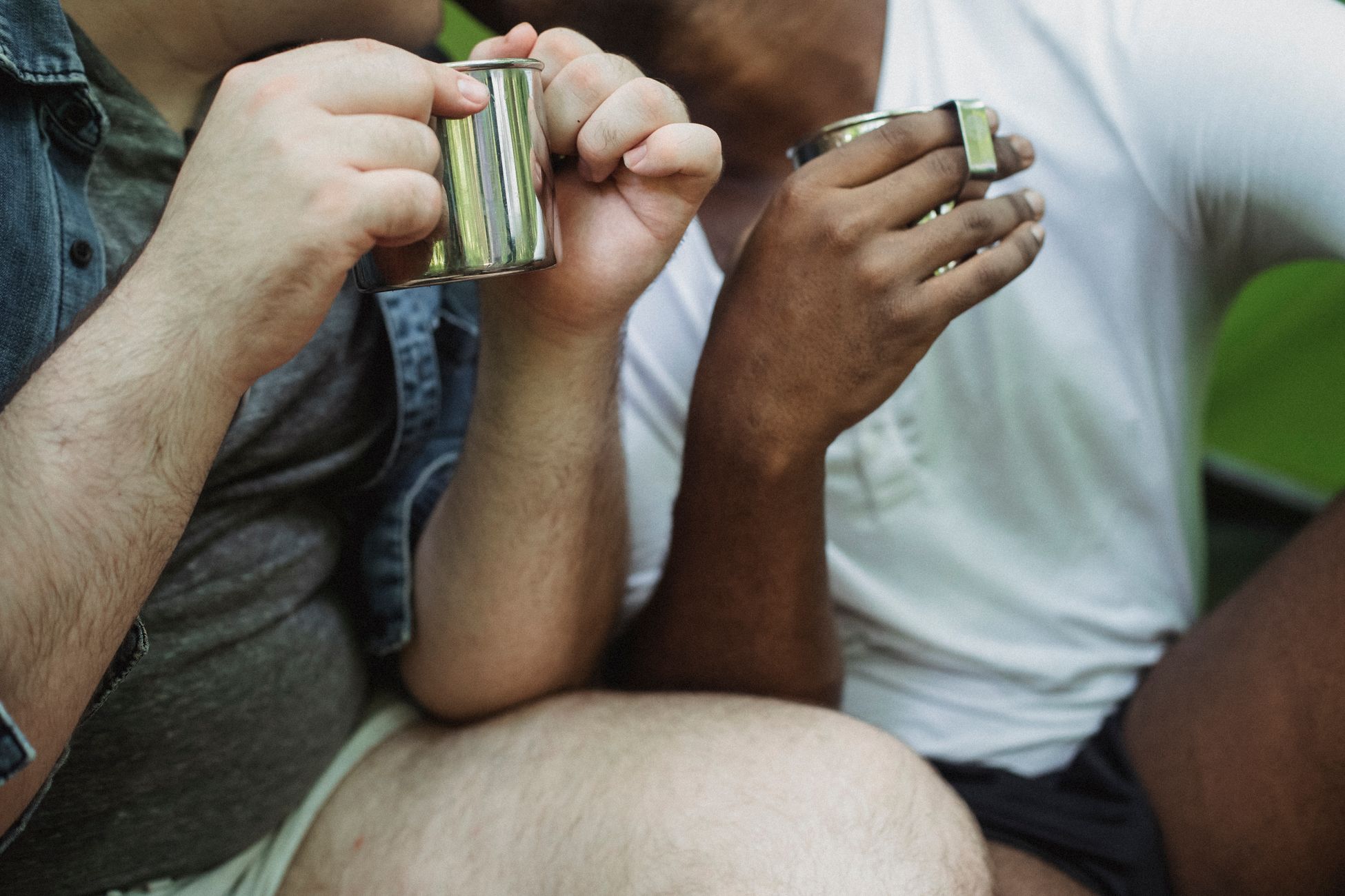 Crop unrecognizable diverse gay couple with camping metal mugs sitting close to each other in daytime