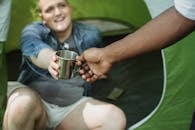 Crop anonymous African American man taking camping metal cup from cheerful friend sitting in tent in daytime