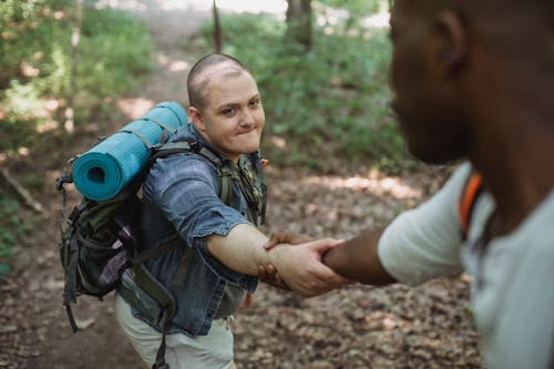Crop anonymous African American traveler helping friend with backpack with hiking equipment to climb up in forest