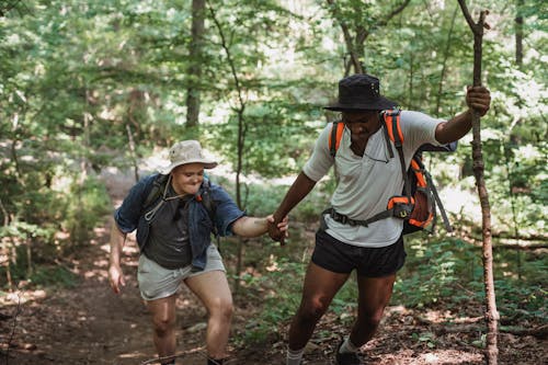 African American traveler in bucket hat with wooden stick helping friend to climb up in forest in daytime