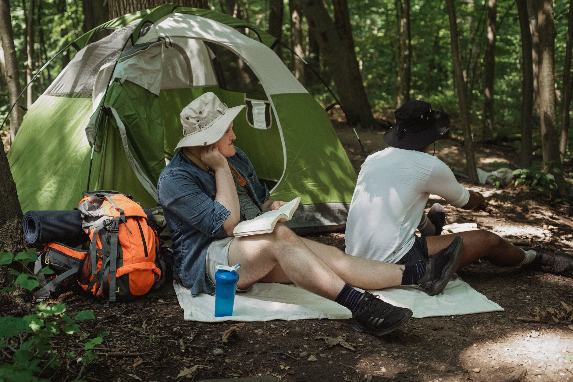 Unrecognizable African American man sitting near green tent next to male friend resting with open book and bottle of water on sunny day