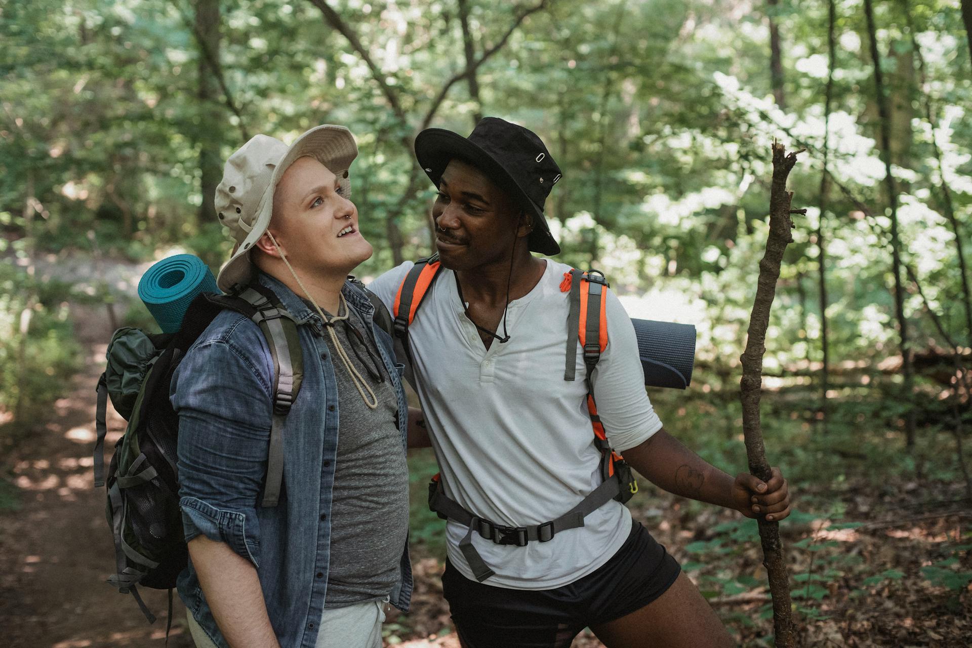 Happy young homosexual couple in tourist hats and with backpacks talking while travelling together in woods during summer vacation