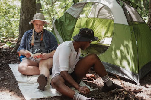 Positive male traveler with retro camera reading interesting book while faceless African American friend sitting near green tent with dry firewood