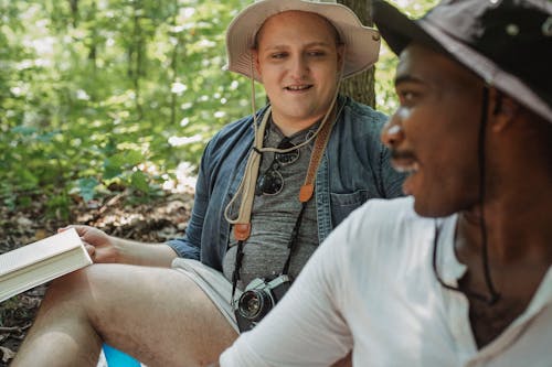 Smiling bearded African American man in black hat looking over shoulder on man while sitting together on blurred background in woods