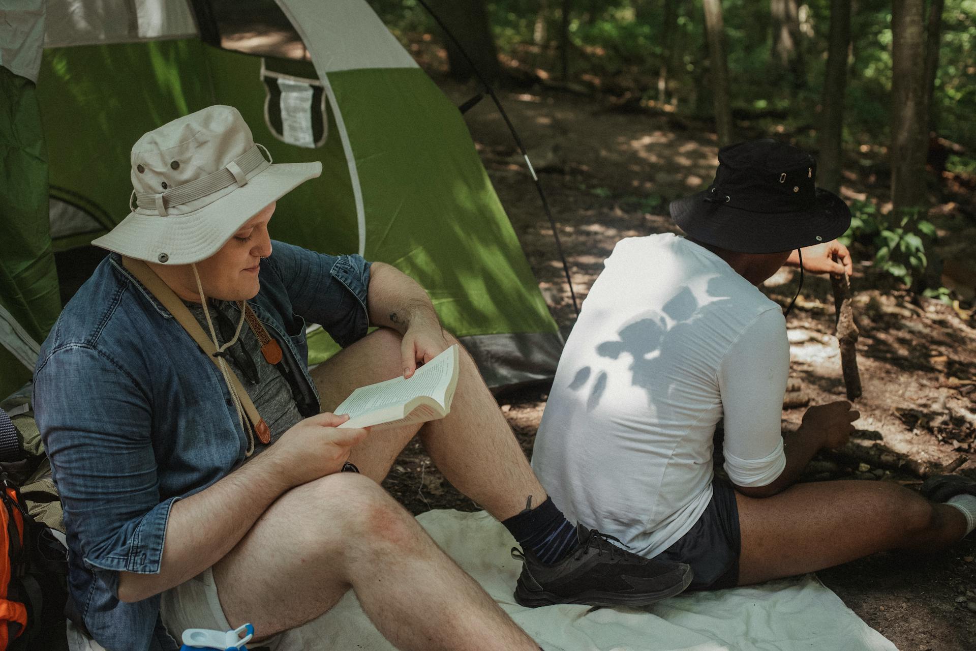 Black male tourist in white shirt with sun reflection of leaves on back sitting with firewood near green tent and his male companion reading book