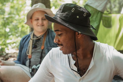 Cheerful African American guy sitting near tent in forest and chatting with friend during hike trip