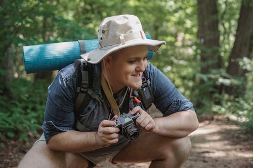 Young smiling male hiker with mat and photo camera squatting on pathway with trees behind while looking away in woods