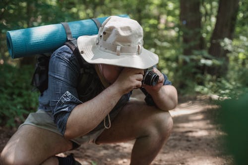 Unrecognizable male tourist with hiking equipment squatting on path while taking photo on camera in summer woods