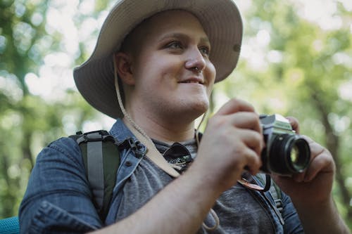 Crop young glad male hiker in panama hat with photo camera looking away in summer forest