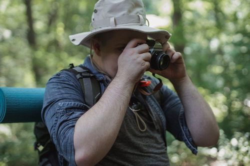 Free Crop anonymous male traveler in panama hat with backpack taking photo on camera in forest Stock Photo