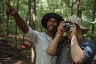 Young black male traveler indicating with finger near anonymous best friend taking photo on camera in woods