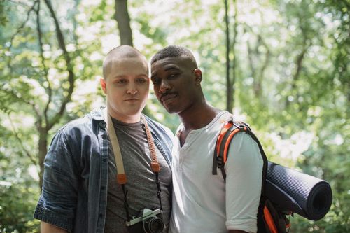 Free Young multiracial male hikers with backpack and photo camera standing in green forest on sunny day and looking at camera Stock Photo