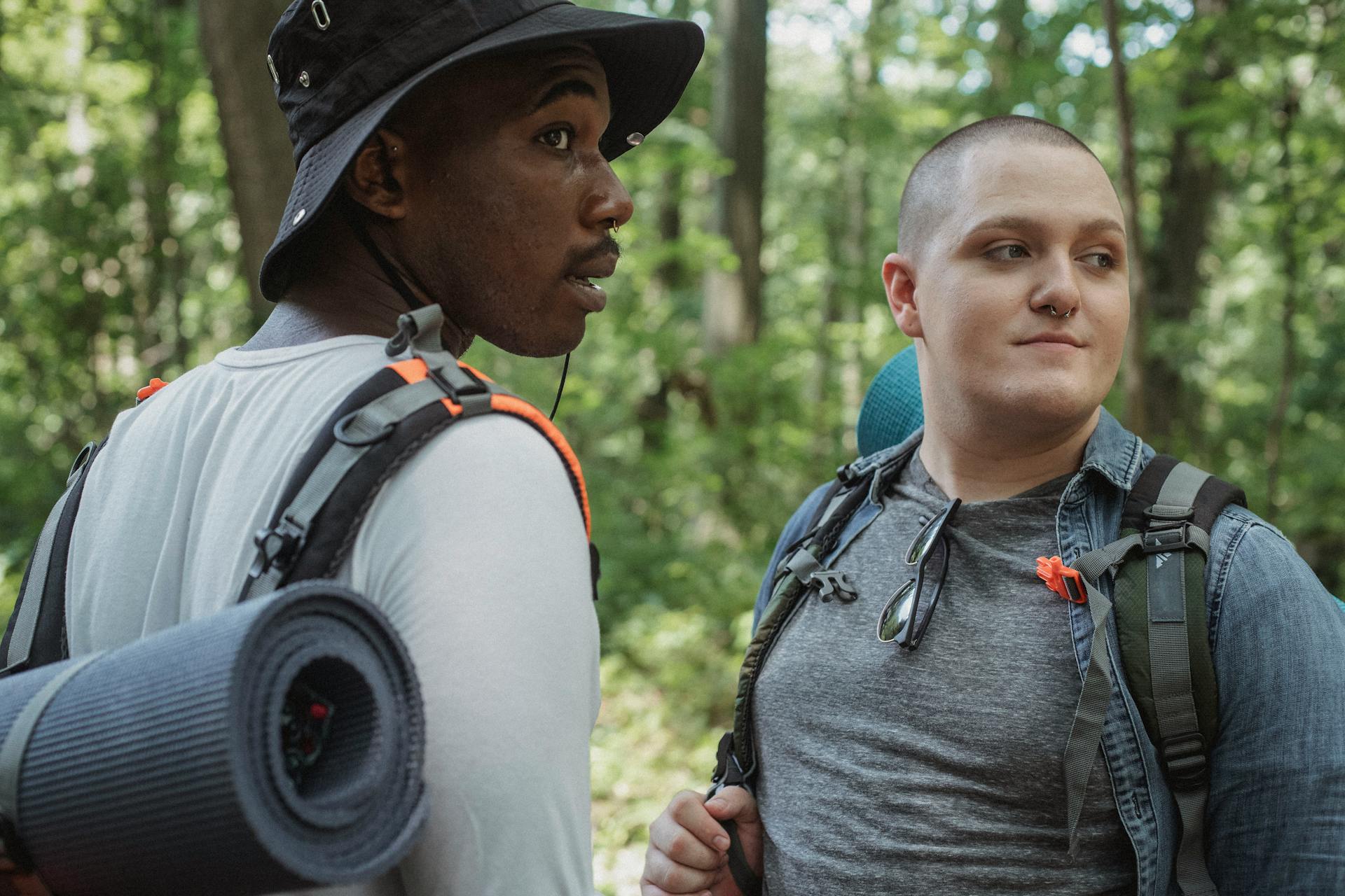 Young diverse male travelers in casual clothes with backpacks and mats resting among green trees in forest during hiking trip in nature