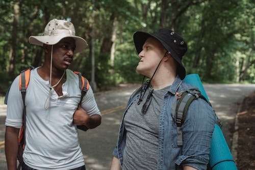Young multiethnic backpackers in casual apparel looking at each other while speaking on countryside roadway near forest