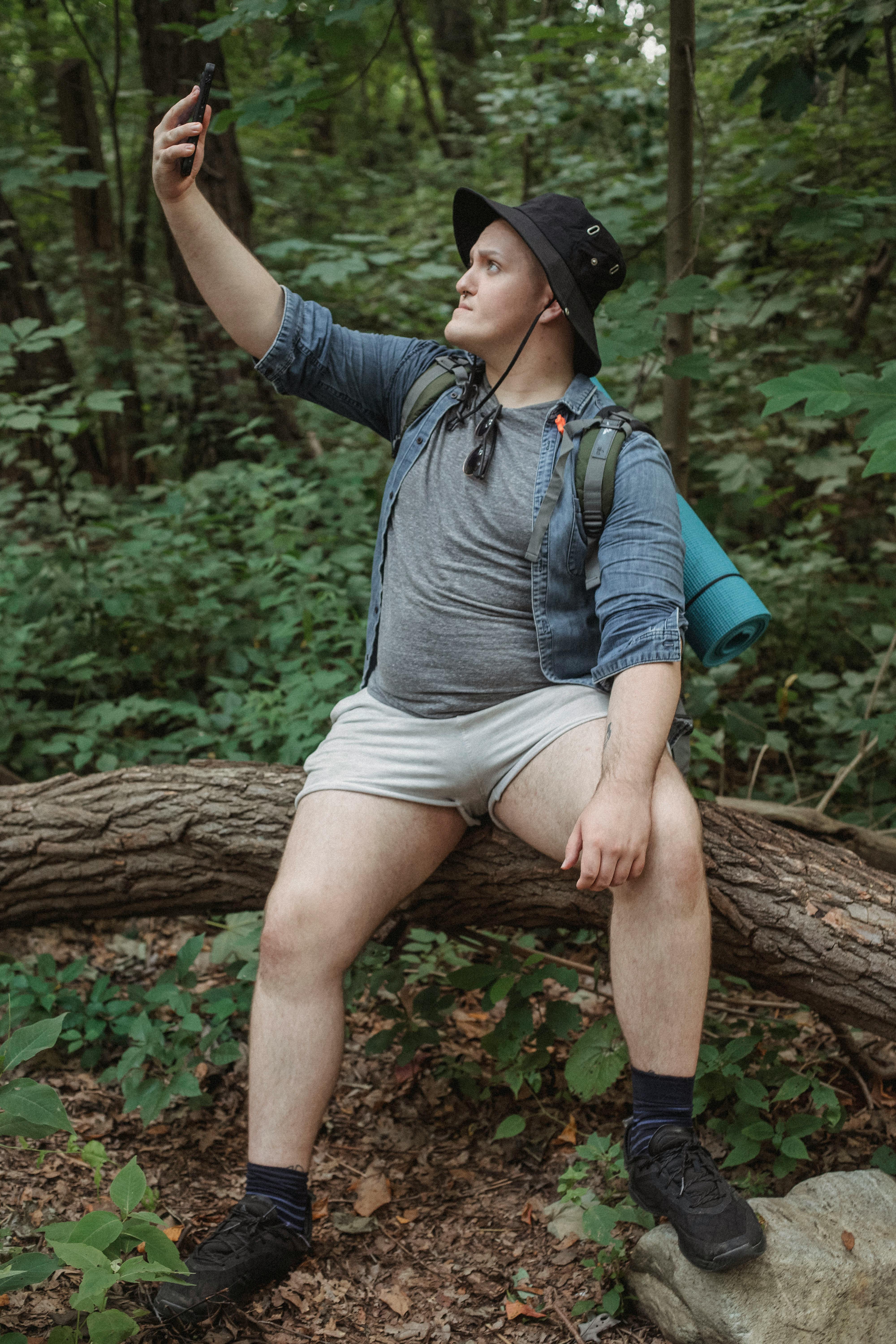 hiker taking selfіe on smɑrtphone whilｅ resting on tｒee trunk