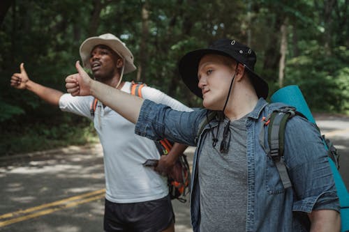 Young multiracial male backpackers in panama hats with hiking equipment standing with thumbs up on roadway against forest and looking away