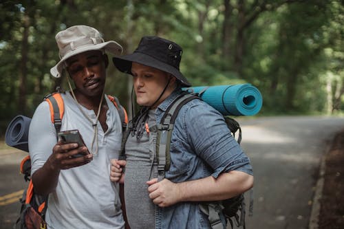 Young multiethnic tourists with hiking equipment surfing internet on cellphone on roadway against trees