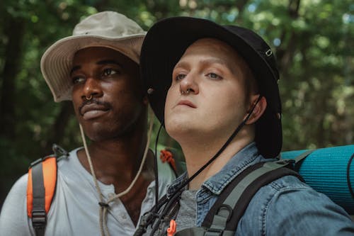 Young homosexual multiethnic men in panama hats with hiking equipment looking away against woods