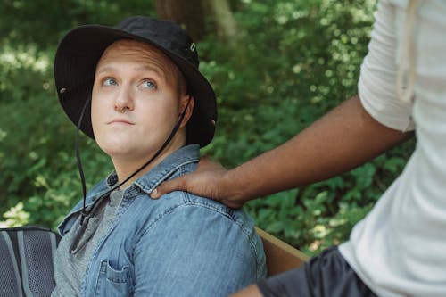 Crop anonymous ethnic male touching shoulder of contemplative beloved in panama hat while resting on bench