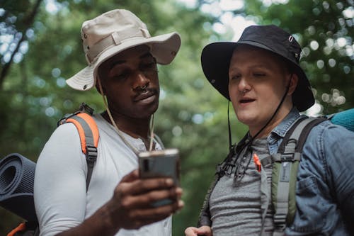 Free Young multiethnic hikers with backpacks watching cellphone against trees while spending time together on weekend Stock Photo