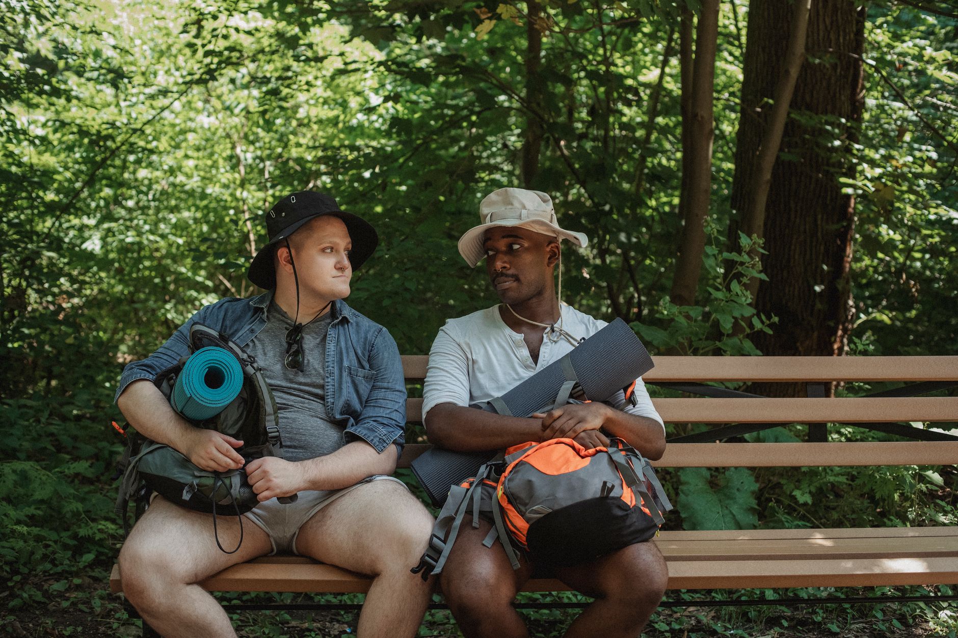 Young multiethnic hikers with rucksacks looking at each other on wooden bench in woods