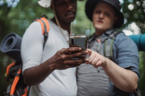 Crop young multiracial male backpackers browsing internet on cellphone while spending time together on weekend in park