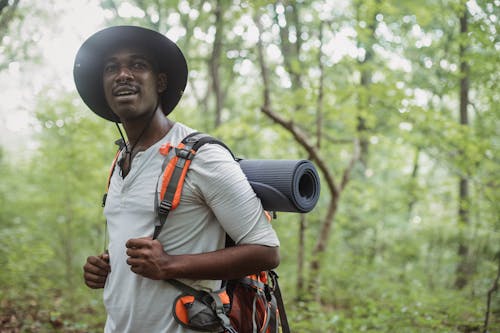 Позитивный черный человек, стоящий с рюкзаком для треккинга в лесу