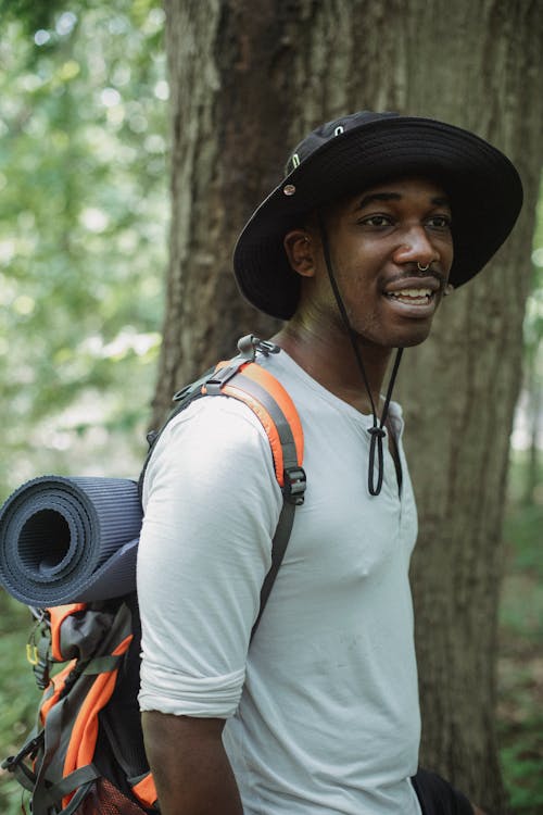 Side view content African American male traveler with trekking equipment and backpack standing in green woods