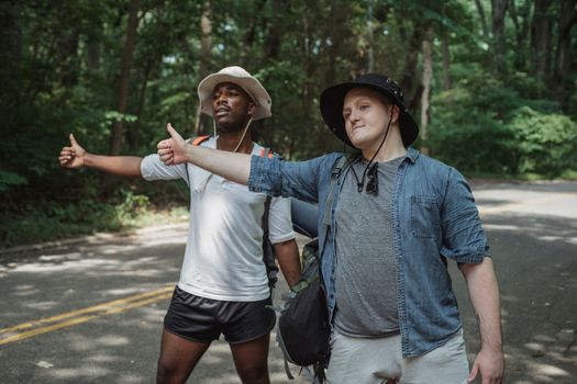 Content multiethnic male hitchhikers in casual outfits and hats with backpacks standing on roadside with thumbs up in lush forest
