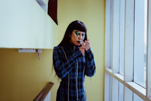 Stylish Asian woman with body art on face cigarette near window