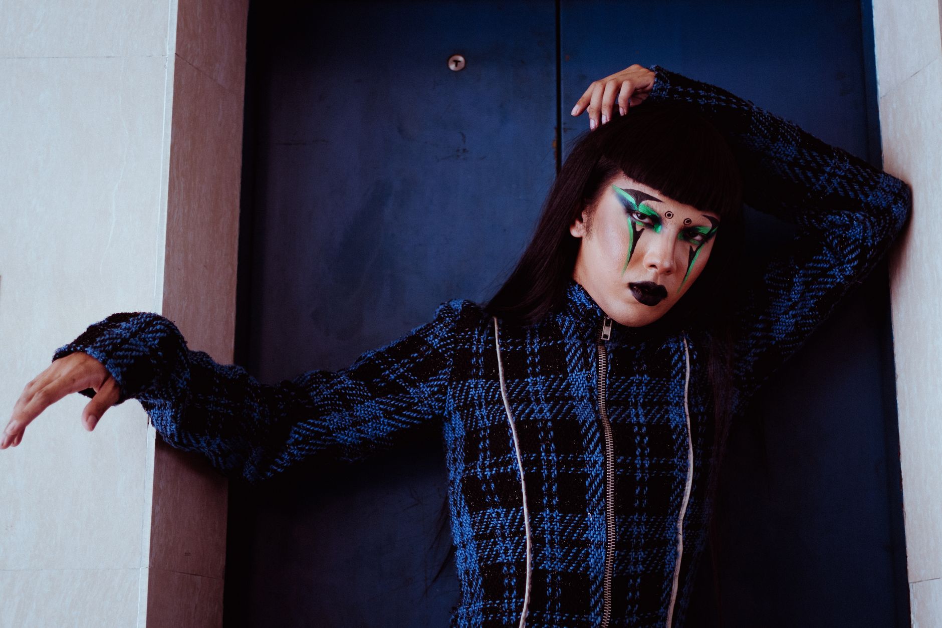 Pensive Asian female with body art on face looking at camera while standing near closed doors of elevator with hand behind head