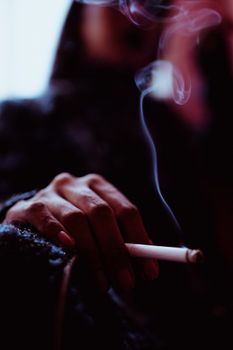 一名模糊的女性吸烟者坐在窗边的房间里，在弱光下放松地抽着烟，吸入烟气