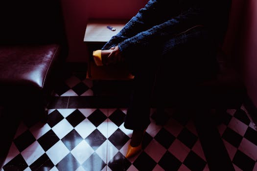 From above of crop anonymous female in trendy footwear and coat sitting on pouffe in room with checkered floor