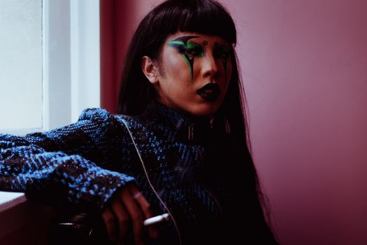 Young ethnic female with extraordinary body art on face looking at camera and smoking cigarette near pink wall