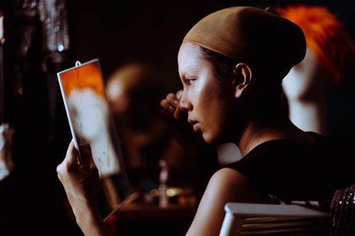 Side view of ethnic female applying cosmetic on face and looking at mirror on blurred background of dressing room