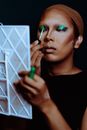 Focused ethnic female applying fluid green and blue eyeshadows while looking at mirror