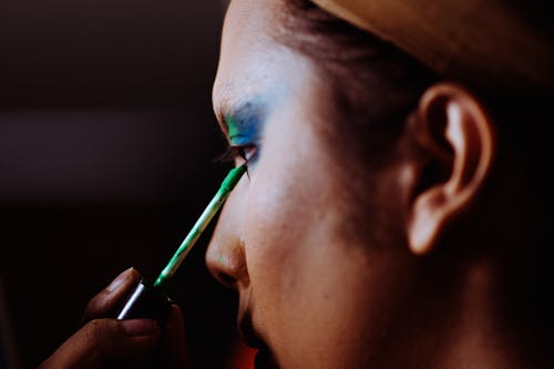 Side view of crop concentrated young ethnic female applying bright makeup on face on blurred background