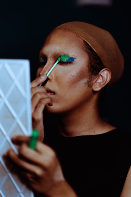 Free Young ethnic actress with mirror applying green eye shadow on eyelid with cosmetic brush Stock Photo