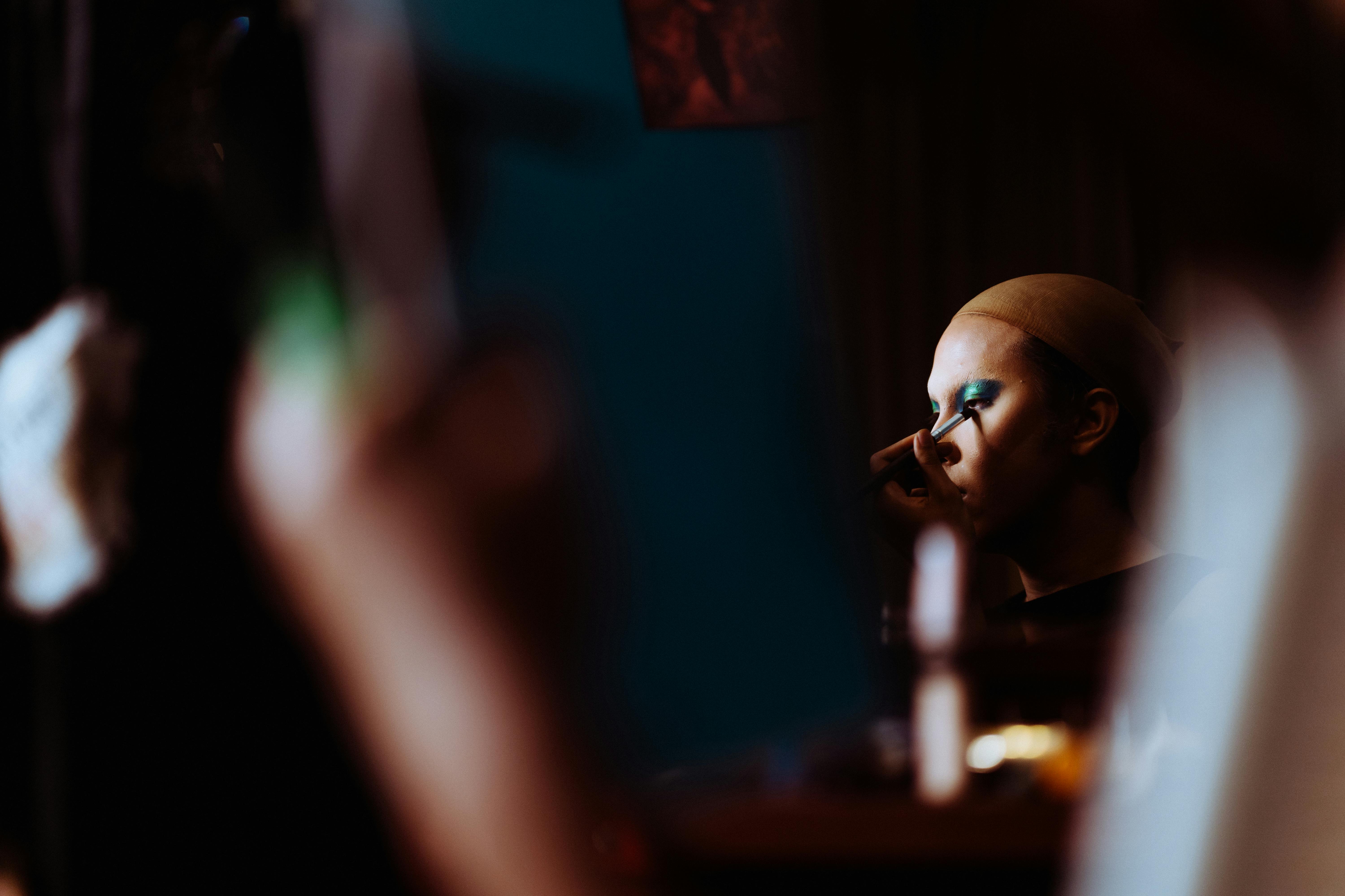 Side view of young theater artist in wig cap applying makeup on face with applicator