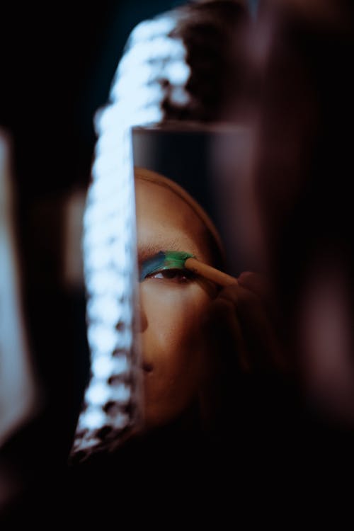 Crop female applying cosmetic on face with foundation and powder and looking at mirror in soft focus