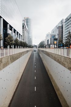 Empty city road lined with skyscrapers and a single moving car under cloudy skies.