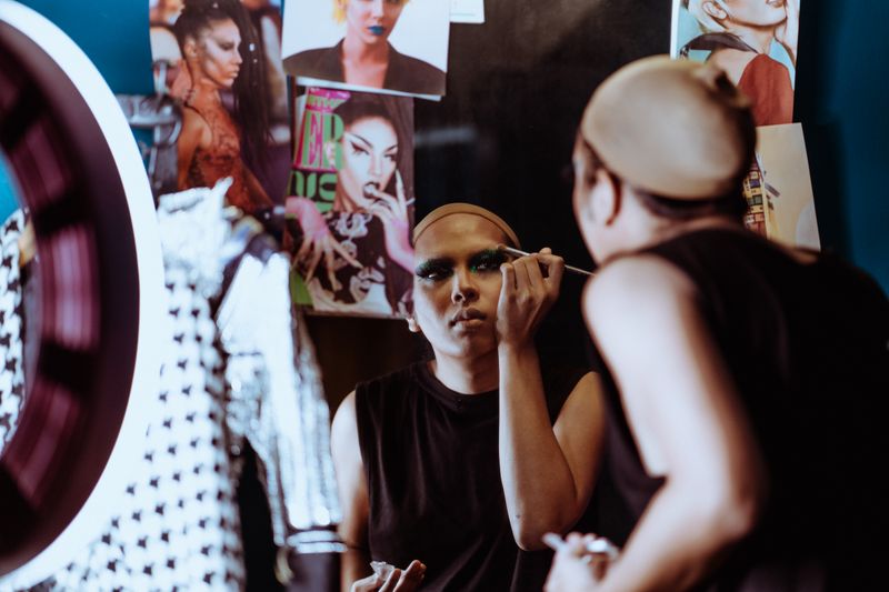 Back view of young attentive ethnic actress looking in mirror with photos while applying makeup with applicator