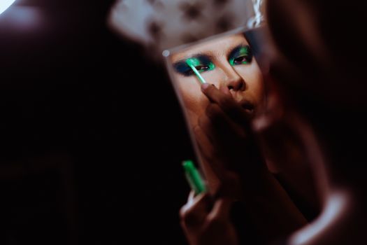 Back view of crop Asian female with mirror applying green eye shadow on eyelid while looking at camera